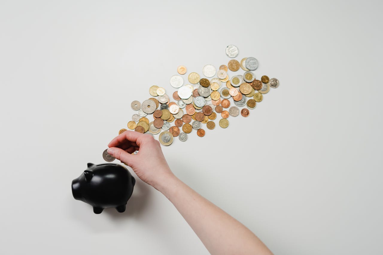 gallery-2 Person placing coin into a black piggy bank with scattered coins, symbolizing savings and finance.